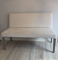 Front view of light beige upholstered settee showing padded seat and backrest with four dark wooden legs on light wood floor.