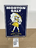 Front photo of rectangular porcelain enameled Morton Salt sign showing navy blue background, yellow-dressed girl with umbrella, 'MORTON SALT' in white text and 'WHEN IT RAINS IT POURS' in yellow text.
