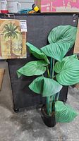 Photo showing faux philodendron plant with large green leaves in black pot and Tuscan palm canvas with beige and green tones hung nearby