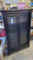 Front view of black media cabinet with closed ribbed glass front doors showing dimensions label on one door.
