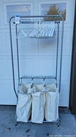 Front view of rolling metal laundry rack with 3 beige canvas hampers and several white plastic hangers hanging from the bar. Top shelf visible above the hangers.