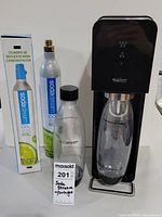 SodaStream machine assembled with clear soda bottle inserted, new CO2 cartridge box and opened cartridge next to it, and an extra soda bottle.