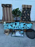 Two handcrafted wood and canvas deck chairs with floral and striped patterns, black metal plant stand holding faux plant, blue decorative metal panel, new black gas grill cover in box, straw bucket basket, and round black metal mesh food cover arranged outside.