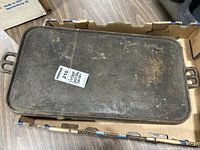 Top-down view of a rectangular heavy cast iron griddle inside a cardboard box, showing flat surface with patina and some rust marks, two handles on each end.