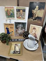 Photo showing full lot items arranged on table including metal signs, framed prints, enamelware pitcher and bowl, light-up letter, and potted ivy plant.