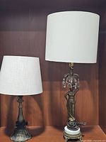 Two antique bronze lamps side by side on a wooden shelf, showing full profile and linen shades
