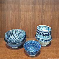 Front view of all nine blue and white ceramic bowls stacked and arranged showing a variety of patterns including checkerboard, floral, leaf, and wave designs.