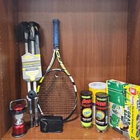 Full view of walking poles, tennis racket, tennis balls, lantern, and grab bags on a shelf