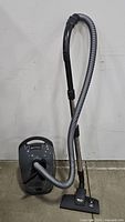 Full view of the Miele AllTeQ grey canister vacuum with hose, wand, and floor nozzle attached. Vacuum is upright against the wall on the floor.