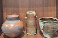 Three ceramic planters/pots on a wooden shelf: large round terracotta pot with textured design on left, tall narrow pottery vase with small ear-handles in center, multicolored glazed pot with rounded rim on right.