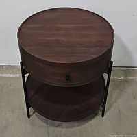Front view of round walnut style nightstand showing the top surface, drawer front with knob, and bottom open shelf supported by black metal frame.