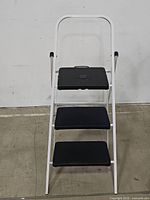 Front view of the 3-step ladder showing the black plastic steps and white metal frame with handle bar.
