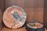 Orange terracotta plate with a black and white bird painted on a branch, on display stand with a tray holding stoneware bowls and a cup.