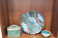 Lot showing the multicolored round platter, lidded ceramic dish, and face bowl displayed on wooden shelf