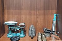 Full view of lot items arranged on wooden shelf: blue kitchen scale with weights, heavy iron, vintage baby shoes, metal press, black weight, and thermometer