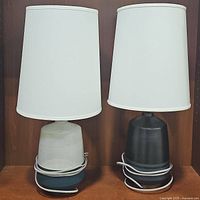 Two vintage vessel style table lamps side by side on a wooden surface, both with white fabric shades. One lamp has a beige base, the other black. Cords coiled around bases.