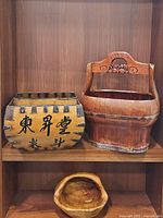 Photo showing an Asian wooden bucket/planter box with black metal reinforcements and Chinese characters, an Asian water or rice wooden bucket with carved handle, and a small wooden carved basket on shelf