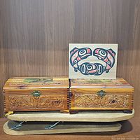 Two ornately carved wooden hinged boxes and the vintage 1980s Haida sliding cedar box with salmon decoration placed on top of a wooden ironing board base