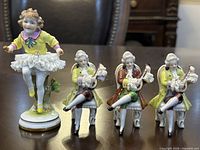 Front view of four porcelain figurines showing three seated Victorian men playing mandolins and one standing girl dancer with lace skirt.