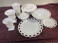Eight-piece set of white milk glass items arranged on a surface, showing various shapes and sizes including plates with reticulated edges, vases, and urns.