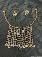 Full view of the gold tone mesh necklace with multicolored crystals and matching drop earrings laid flat on black fabric.