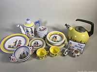 Overview of lot showing yellow enamel kettle, coffee pot, tin/canister, plates, cups, creamer, bowls, decorative tile. All hand-painted Henriot Quimper pottery with Breton woman motifs.