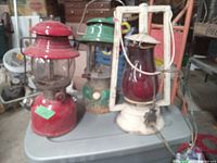 Overall photo showing three vintage lanterns placed on a plastic surface: red Coleman, green Coleman, and a white metal lantern with red glass globe and power cord.