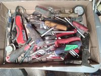 Box filled with assorted used hand tools including screwdrivers, pliers, a manual hand drill, wrenches, and a pressure gauge.