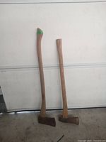 Photo showing two axes standing upright against a white background, both with wooden handles and metal heads