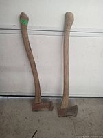 Two used axes leaning against a white wall, showing worn wooden handles and metal heads in used condition.