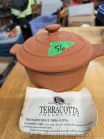 Photo of a small terracotta pot with lid on a wooden surface, with a green sticker labeled '36' and an informational paper about terracotta properties underneath.