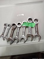 Photo of 7 stubby ratcheting wrenches aligned side by side on a flat surface. The brand name Sterling is seen on the surface beneath the tools. A green tape label marked with the number '5' is on the upper side of one wrench.