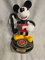 Front view of the Disney Mickey Mouse rotary phone, showing Mickey holding the handset and the rotary dial with numbered buttons on base.