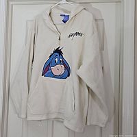 Front view of off-white fleece hoodie with large Eeyore face printed on front pouch pocket, "Eeyore" word on chest, hanging on white hanger against door