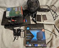 A photo showing a black Gateway desktop tower with AMD and NVIDIA stickers, a small Dell desktop tower, a collection of DVDs including Family Guy 2010, a small black oscillating fan, two white smartphones, a clock radio, a remote, assorted cables, and a boxed Clear TV antenna.