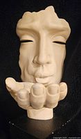 Close-up front view of sculpture showing detailed human face with eyes closed and lips puckered as if blowing a kiss, hand extended below lips.