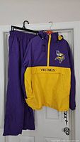 Full view of the Minnesota Vikings tracksuit showing jacket and pants hanging on hangers against a door.