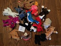 Full view of 12 assorted Beanie Babies laid out on wooden floor, showing various animals including bears, birds, and others, several with original Ty tags.