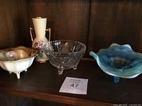Four items displayed on wood shelf: cream footed candy dish with brown rim, German porcelain urn vase with violet flower design and gold handles, clear cut glass candy bowl with faceted pattern and three ball feet, blue opalescent ruffled glass dish with handles and three feet.