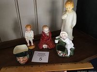 Five children figurines displayed with a ruler for scale showing tallest is approximately 8 inches tall. Includes two seated figurines, a standing figurine, a girl leaning on a green base, and a small character head mug.