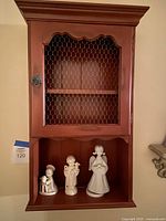 Front view of burnt red wooden hanging curio cabinet with wire mesh door and open shelf below, showing three porcelain figurines arranged underneath.