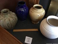 Set of four decorative pottery vases on a dark surface, showing the variety in color and texture including a greenish taupe, deep blue, mustard, and cream colors with ribbed designs.