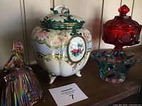 Four decorative pieces shown together: ceramic urn with floral and gold detail, iridescent colonial girl figurine, red glass candy dish, and a decorative glass bowl with colored leaf-like patterns.
