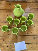 Top-down view of the lime green pitcher with 8 matching mugs arranged around it on wood surface.