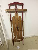 Full view of the Flexible Flyer III sled standing upright against a wall, showing the wooden slats, red metal runners, and rope handle.