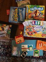 Photo showing various board games stacked including Trouble, Scrabble Junior, Life, Flinch, Pit, and additional card games scattered with game components.