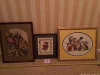 Three framed cross stitch pieces leaning against a wall, showing the full lot contents including cottage, alphabet bear, and Snow White scenes