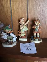 Three Hummel figurines displayed on a shelf in front of a wood-paneled wall, including the boy with bunnies, girl with flower, and girl with cat.