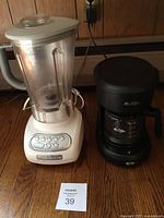 Photo showing both KitchenAid blender and Mr. Coffee coffee maker placed side by side on wooden surface against wood panel wall.