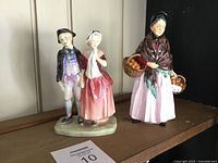 View of two Royal Doulton figurines on display: one courting couple, one elderly woman with baskets of oranges.